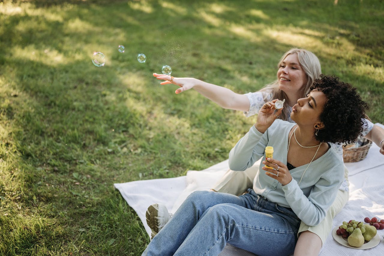 Two women enjoying an outdoor picnic, blowing bubbles on a sunny day.